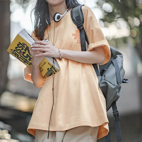 Person wearing a beige t-shirt with text, holding a book, and standing outdoors.